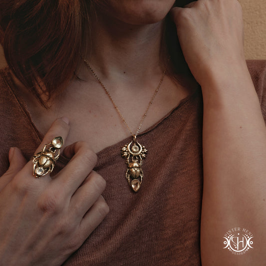 Close-up of a person wearing a gold scarab necklace and ring, with a blurred background.
