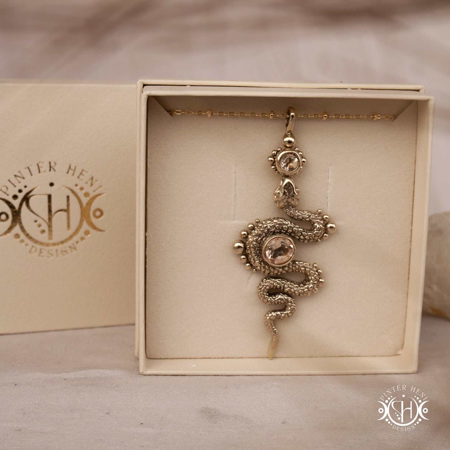 Decorative gold snake pendant in a box with 'Pinter Heni Design' branding.
