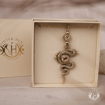 Decorative gold snake pendant in a box with 'Pinter Heni Design' branding.