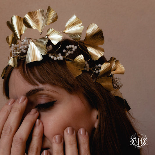 Woman wearing a decorative gold headpiece with leaves and berries, holding her face.