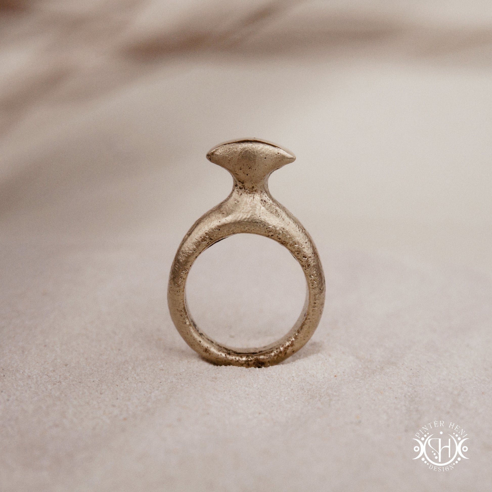 Glyph Ring handmade sandcast brass ring with curved sculptural top and ancient symbolic character, on sand.