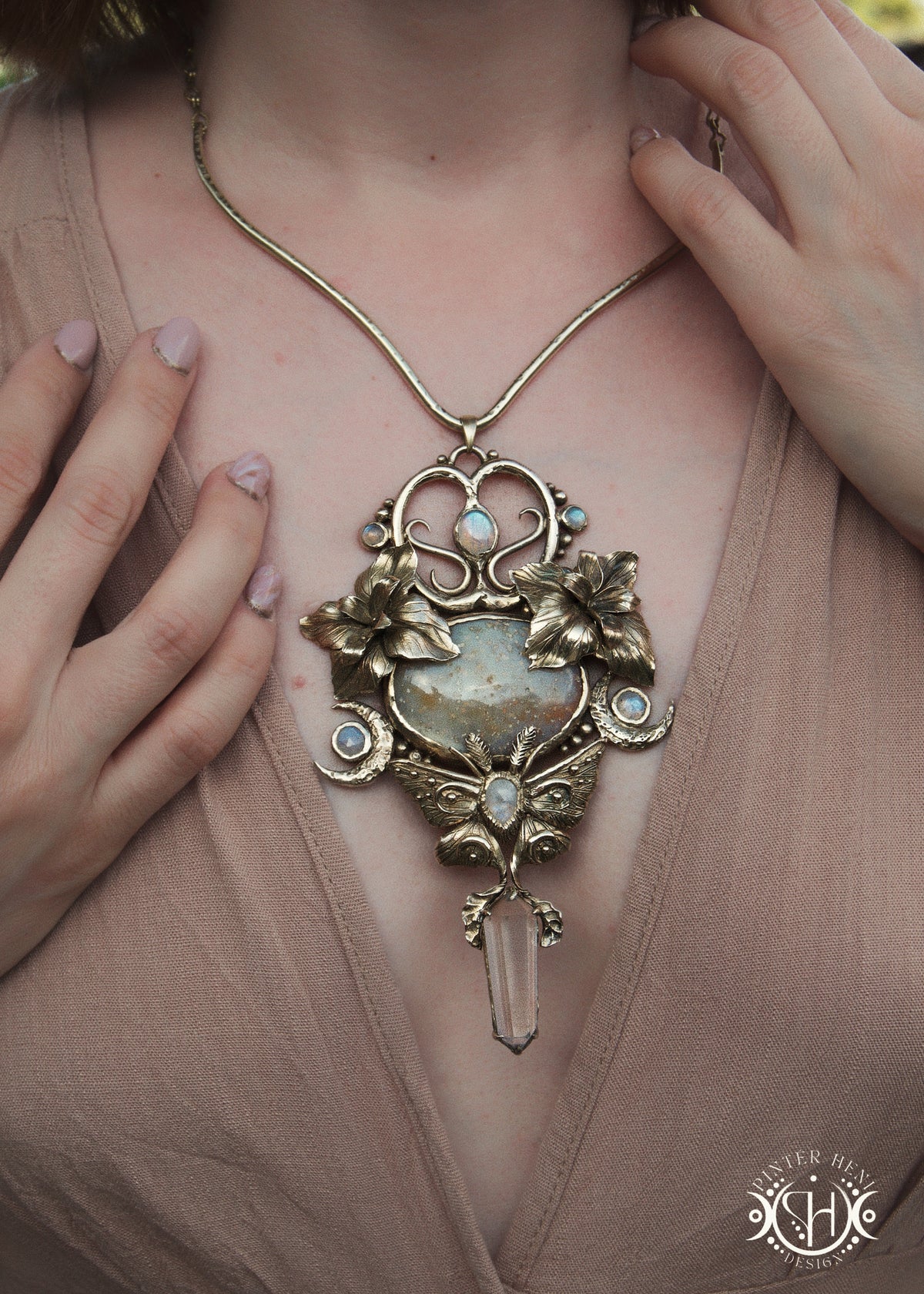 A close-up lifestyle shot of a model wearing a large, ornate vintage-style pendant necklace. The necklace features a gold-toned filigree frame with floral and mothdetails, a central big ocean jasper, small light blue moonstones, and a clear quartz crystal at the bottom.