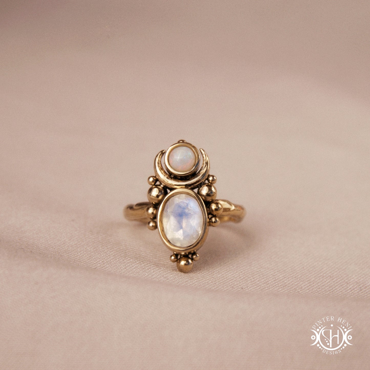Hand-carved Nyx ring with opal and moonstone in brass, sculptural crescent design, on beige textile.
