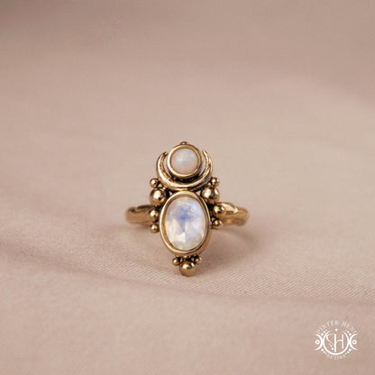 Hand-carved Nyx ring with opal and moonstone in brass, sculptural crescent design, on beige textile.
