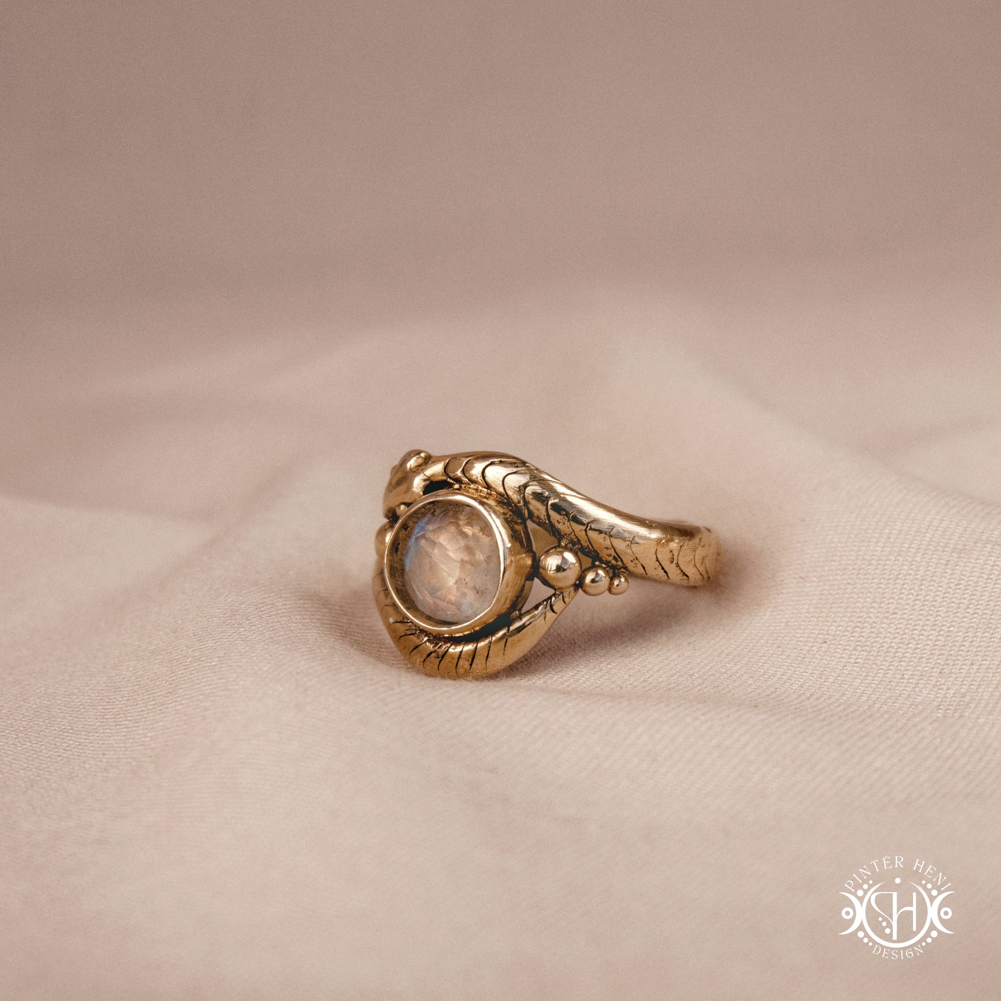 Handmade serpent ring set with moonstone, sculptural wax carved design in brass, silver, or solid gold, side view, on a beige textil.