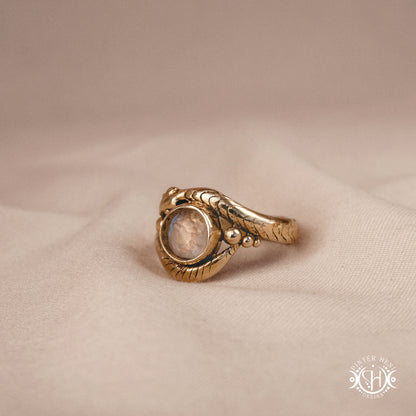 Handmade serpent ring set with moonstone, sculptural wax carved design in brass, silver, or solid gold, side view, on a beige textil.