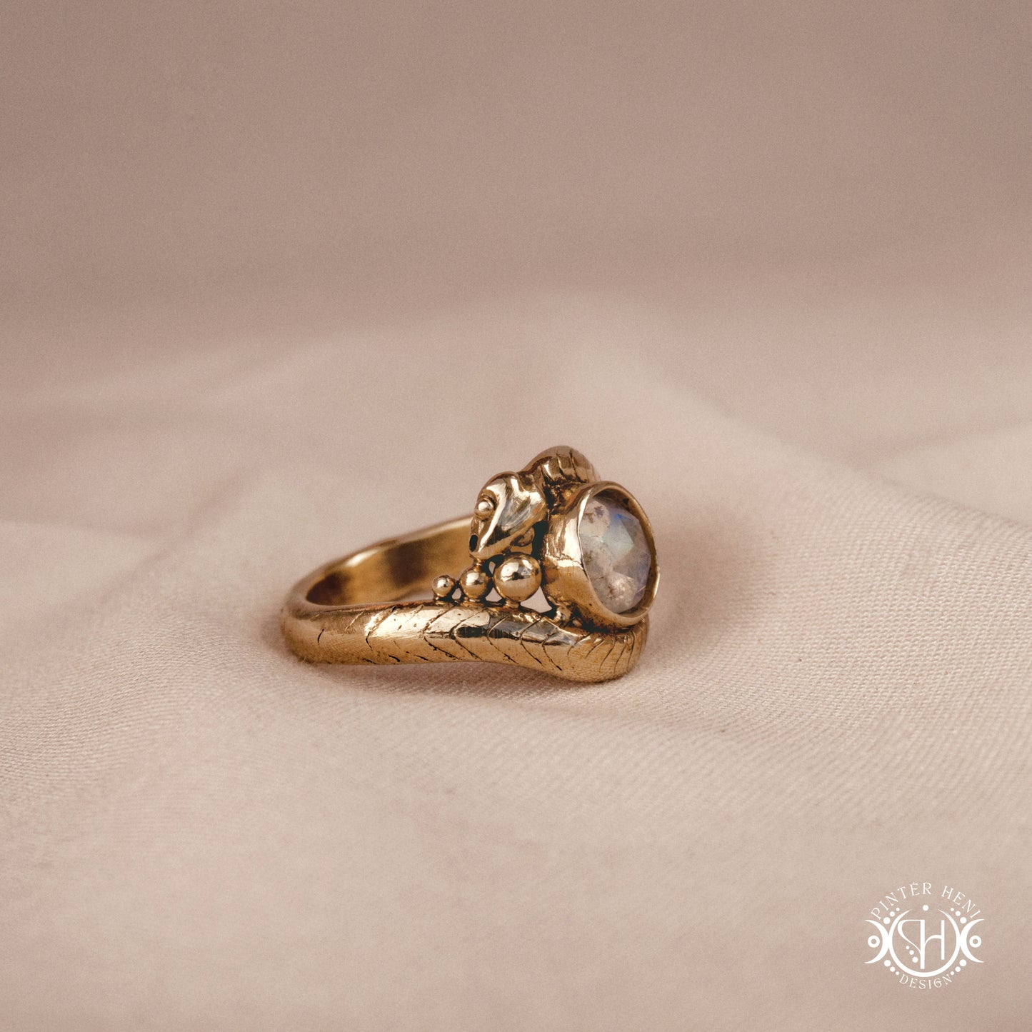 Handmade serpent ring set with moonstone, sculptural wax carved design in brass, silver, or solid gold, side view, on a beige textil.