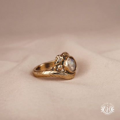 Handmade serpent ring set with moonstone, sculptural wax carved design in brass, silver, or solid gold, side view, on a beige textil.