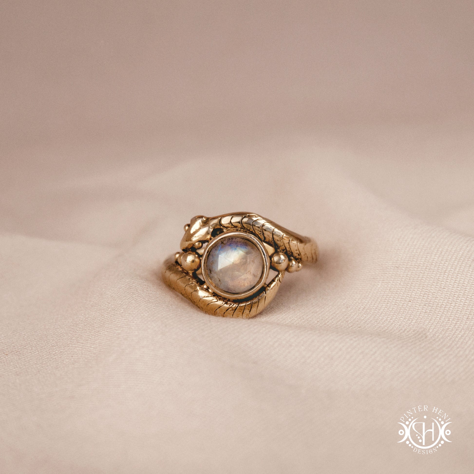 Handmade serpent ring set with moonstone, sculptural wax carved design in brass, silver, or solid gold, close up, on a beige textil.