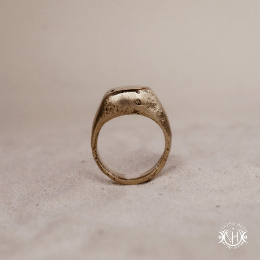Sigil Ring handmade sandcast brass signet-style ring with raw surface and ancient-inspired sculptural form, in sand.